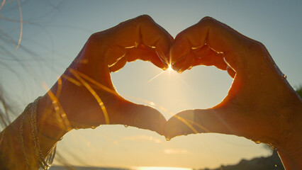 CLOSE UP, LENS FLARE, SILHOUETTE: Woman with bracelets forms a heart shape with hands round the bright sun above sea. Warm golden light spreads across the beach and sky in a summer vacation moment.