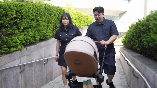 Young couple pushes stroller on city sidewalk. They walk calmly, hands together on handle. Green hedges line concrete path beside them. Sunlight glows softly on stone steps and railings