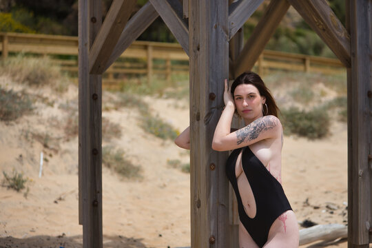 Young, beautiful, Hispanic woman, very tattooed, wearing a black trikini, looking at the camera calmly and relaxed, leaning on the lifeguard tower pole at the beach. Concept of vacation, summer, sun.