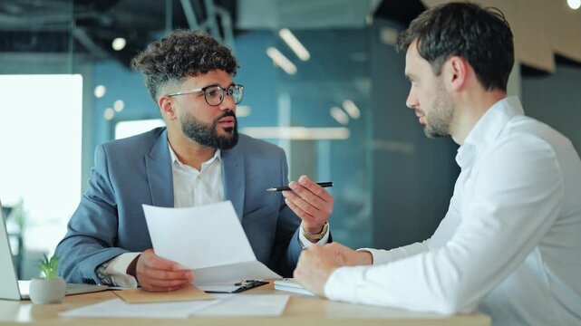 Middle Eastern candidate in suit talking and holding resume during interview with Caucasian employer. Diverse professional men discussing application document or contract at office desk.