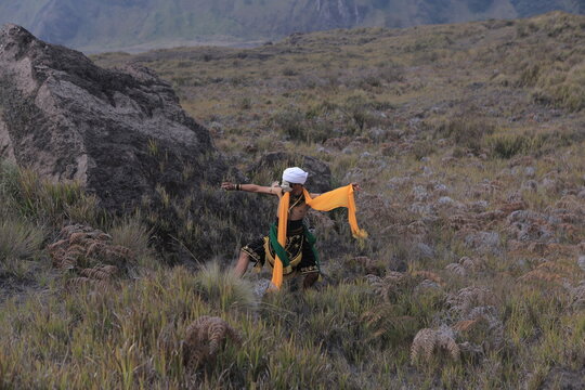Traditional Javanese mask dancer performing outdoors amidst large volcanic rocks and mountains.
