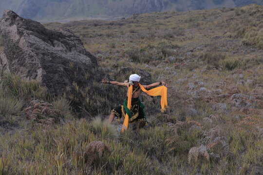 Traditional Javanese mask dancer performing outdoors amidst large volcanic rocks and mountains.