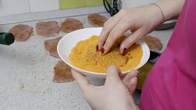 Female hands coat a thin pork cutlet in breadcrumbs after dipping it in an egg wash. She places the prepared schnitzel into a hot frying pan with other cutlets.