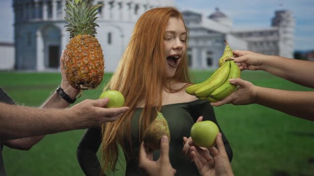 Redhead woman smiling with finger raised as multiple hands offer pineapple, pear and bananas in front of a historic building; playful joy.