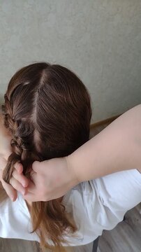 Woman braiding her own long brown wavy hair into a French braid or fishtail plait at home. Close-up over-the-shoulder rear view. Morning beauty routine, hair care, and styling concept video.