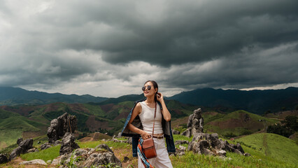 A woman in sunglasses stands on a rocky hilltop surrounded by lush green mountains under dark, dramatic storm clouds. © waragron