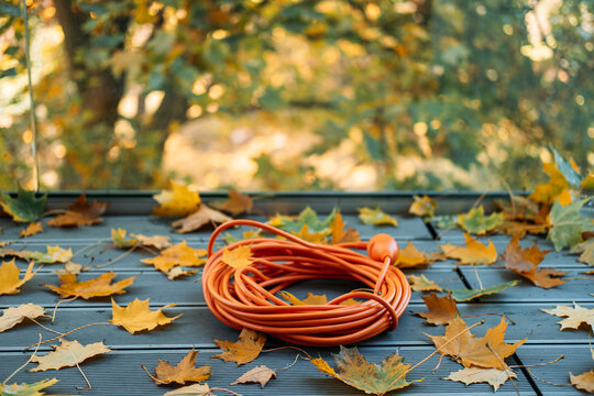 orange coiled extension cord lies on gray ribbed wooden decking next to scattered yellow autumn maple leaves. Outdoor cable insulation, waterproof electrical equipment, extension cord safety.