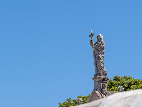 Historische Steinfigur mit Kreuz auf Kapellendach vor blauem Himmel