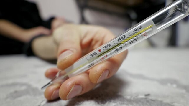 Person's hand holds a glass mercury thermometer showing a high body temperature indicating a fever. Close-up shot with a rack focus from a sick person lying on a carpet in the background.