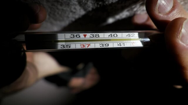 Hands hold a classic mercury thermometer indicating a high fever, with the reading over 39 degrees Celsius. A single light source illuminates the device in a dark, dramatic setting.