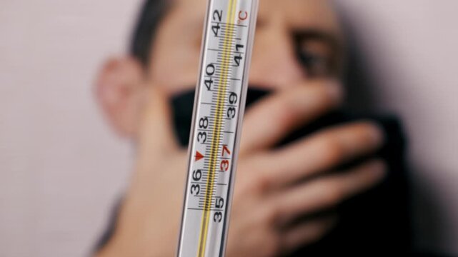 Mercury thermometer shows a rising body temperature, indicating a high fever. Out of focus sick man coughs in the background, creating a shallow depth of field effect.