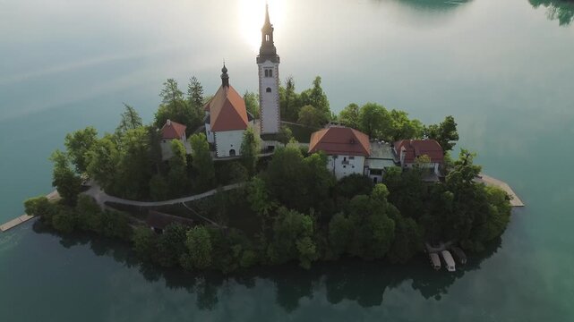 Aerial of the Assumption of Mary Church tower on Bled Island, Slovenia. Beautiful cinematic drone footage of the historic church with the sun reflecting on the turquoise lake water in the background.