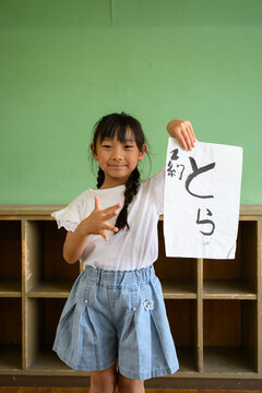 教室の後ろで笑顔で「とら」の習字作品を高く掲げる女子小学生