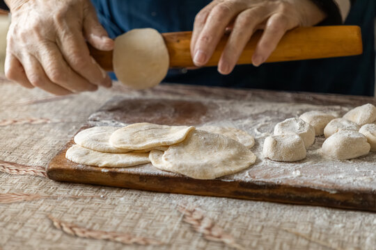 Close up of a person using a wooden rolling pin to flatten dough circles on a floured board, capturing the craft of traditional homemade pastry preparation in a rustic kitchen