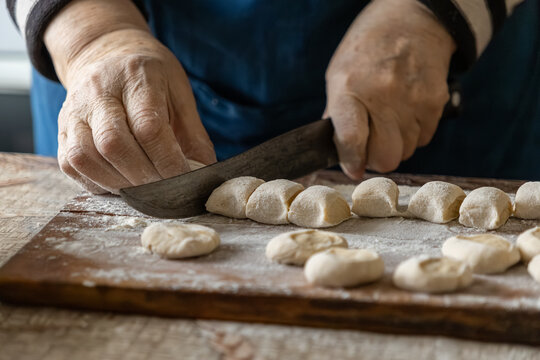 Elderly person hands cutting fresh dough pieces with a sharp knife on a wooden board, showcasing traditional culinary preparation and the authentic art of homemade dumpling making