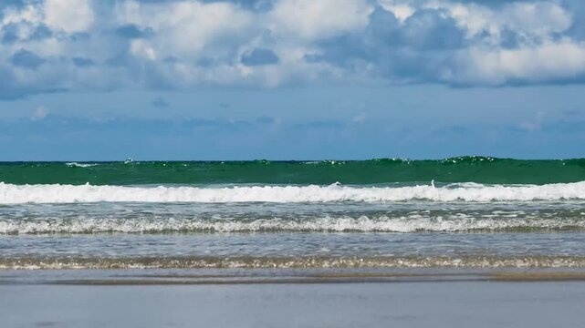 Brechende, t&uuml;rkisfarbene Meereswellen mit wei&szlig;er Gischt rollen unter blauem Himmel sanft an den flachen Sandstrand.