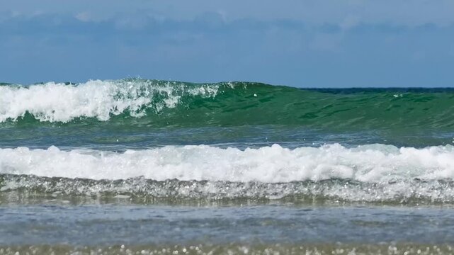Brechende, t&uuml;rkisfarbene Meereswellen mit wei&szlig;er Gischt rollen unter blauem Himmel sanft an den flachen Sandstrand.