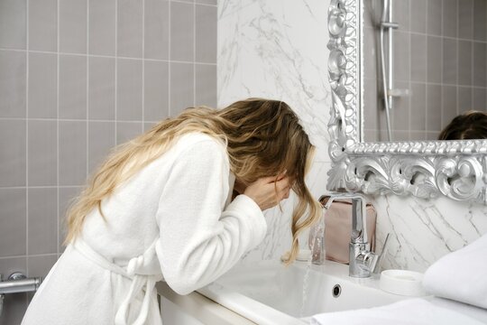 A woman in a white bathrobe leans over a sink to wash her face in a stylish bathroom