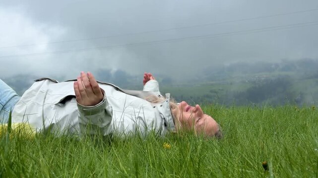 Middle aged woman lying on green grass with arms outstretched and breathing deeply while enjoying mountain landscape. Fog covers valley and village near Gubalowka in Tatra Mountains, Poland.