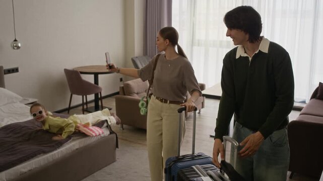 Wide shot of Caucasian couple with child checking into hotel room, woman recording video on smartphone while girl jumping on bed. Perfect for family travel promotions and hotel booking advertisements