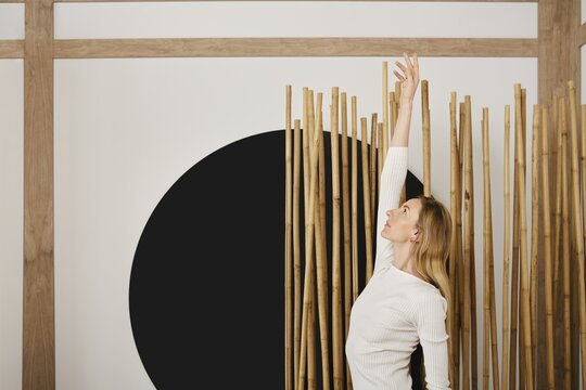 A woman stands in a room reaching her arm upward. She is positioned near bamboo sticks that are grouped together. A large black circle is painted on the white wall behind her