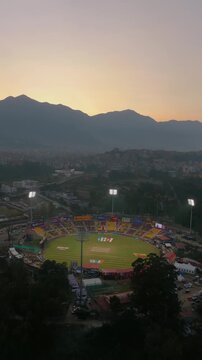 vertical, 9:16, orbiting drone shot circles a live cricket match at TIA Ground in Nepal, showing active players, lit stadium floodlights, and spectators during a colorful evening sunset twilight.
