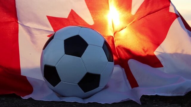 A soccer ball rests on the ground with a flowing Canadian flag behind it. Golden sunlight shines through the flag, highlighting its maple leaf