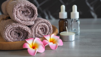 Luxury spa still life with essential oil dropper bottles (amber and clear glass), a scented candle, soft pink mauve towels tied with rope, and pink frangipani flowers on a light gray wooden table. Pre © kanjana