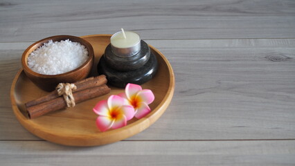 Minimalist spa still life with a soft mauve pink towel tied with rope, a scented candle on black massage stones, and white frangipani flowers in a circular wooden tray. Front view on a light gray wood © kanjana