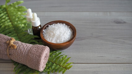 Premium minimalist spa still life with white bath salt in a wooden bowl, mauve rolled towel, cosmetic dropper bottles, and white frangipani flowers arranged in a tray with a green fern leaf. Set on a  © kanjana