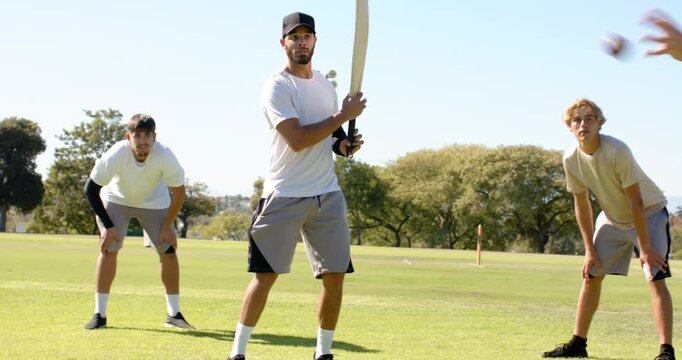 Practicing diverse male teammates playing cricket on park pitch, with bat, ball, stumps