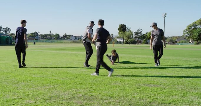 Man tossing red ball to teenagers catching, diving, returning during drill on cricket field