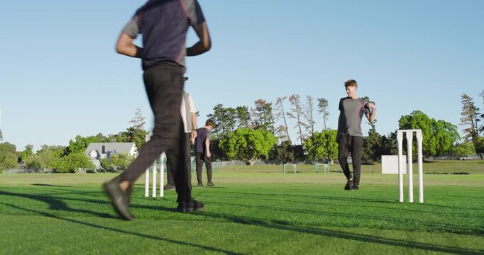 Diverse male friends bowling underarm red ball at stumps on field, rotating, newcomer arrival