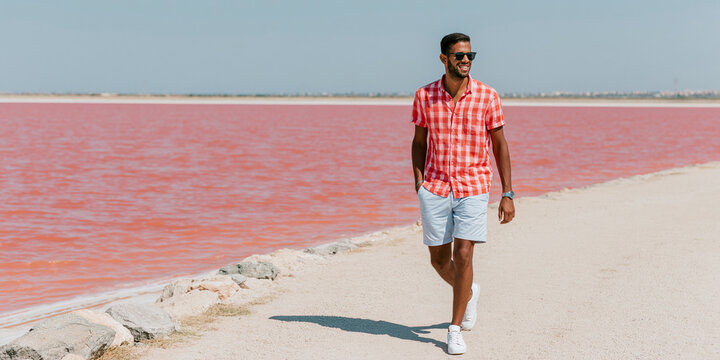 A man in a stylish checkered shirt and shorts walks along a paved path beside a striking pink lake under a clear blue sky. He wears sunglasses and a watch.