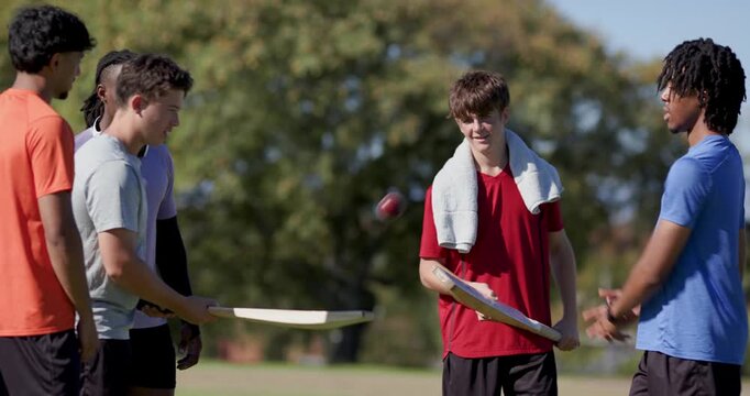 Diverse male friends in park tossing red ball, guiding teen in red practicing bat control