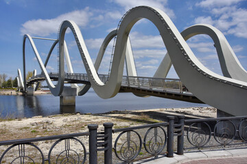 A modern wavy pedestrian bridge spans a calm river at the waterfront. Contemporary architectural arcs create a scenic riverside landmark.