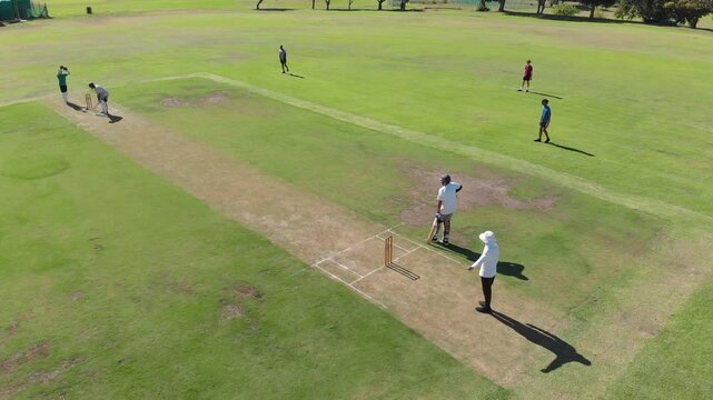 Bowler bowling ball on pitch causing batsman defending as fielder bails