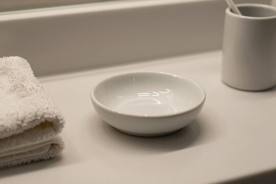 A ceramic soap dish sits on a bathroom countertop. The white empty saucer rests near a folded towel. The porcelain dish is placed on a clean surface.