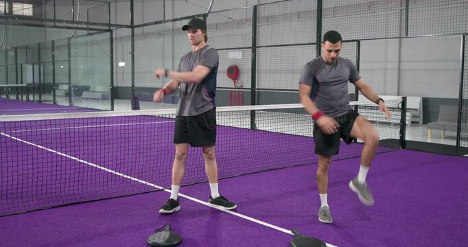 Two male athletes warming up being guided by partner in stretching beside net on purple padel court