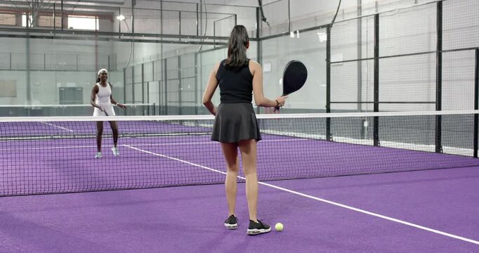 Diverse female friends serving and volleying padel ball using paddles on tennis court