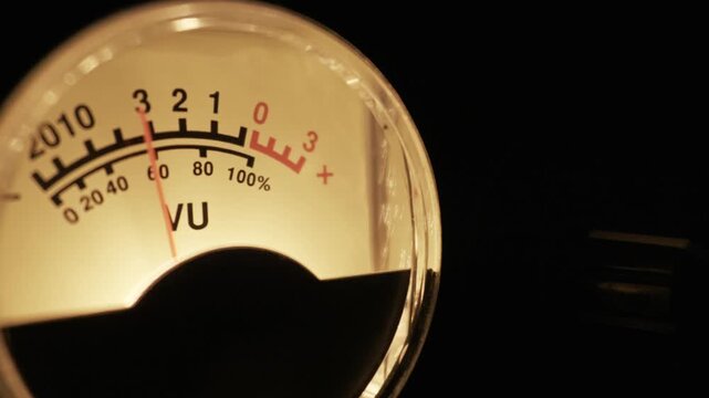 A backlit analog VU meter needle fluctuates while measuring a sound signal level. Concept of audio engineering, recording studio work, music production, and sound mixing.