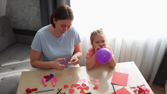 Young mother and little daughter creating paper crafts and inflating balloons together at home. Early childhood development, creativity, and family bonding concept. Top view