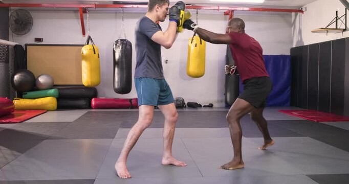 African american coach shifting focus pads and guiding male student's footwork and strikes in gym