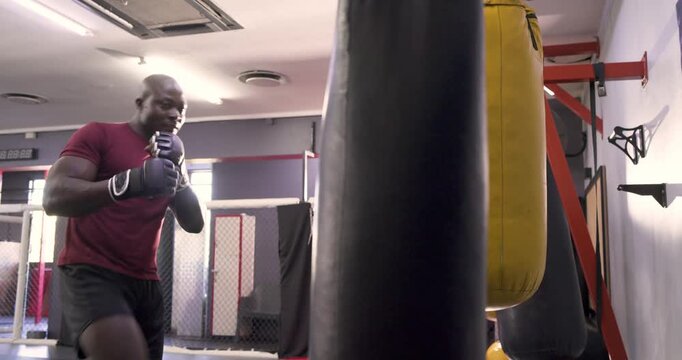 Black boxer performing footwork punching heavy bag inside boxing ring while white man
