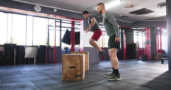 Diverse male partners alternating jumps on boxes in crossfit gym after landing building strength