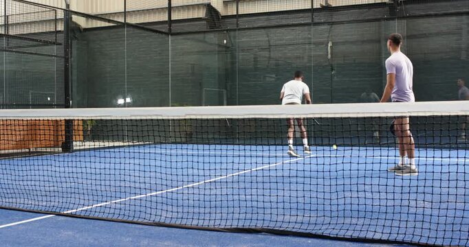 Diverse male players practicing volley drill after ball feed on padel court, lunging, shuffling