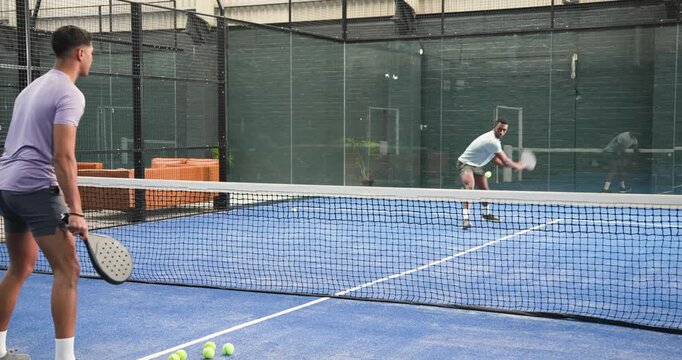 Diverse male training partners feeding padel balls with paddles, volleying on padel court