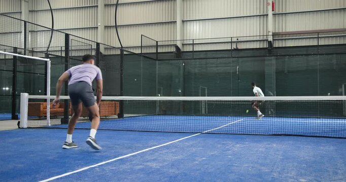 With ball arcing male padel player lunging, volleying on turf court then smashing wall