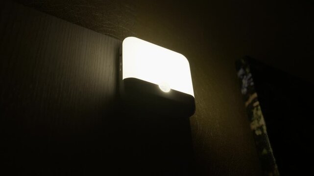 A modern LED night light with a motion sensor is turned on, casting a warm glow on a wall in a dark room. Concept of smart home technology, home automation, safety, security, and energy efficiency.