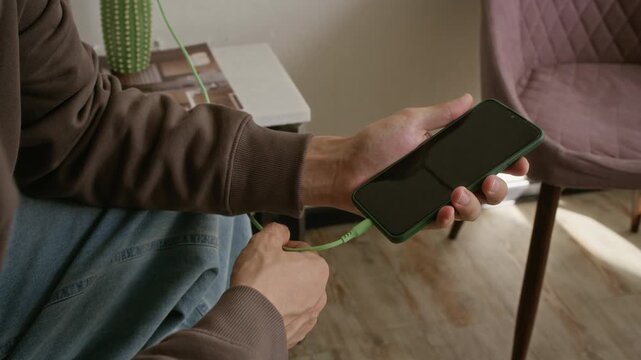 Cropped shot of young man plugging green charger cable into smartphone. Footage is perfect for mobile technology, battery power, and device connectivity marketing campaigns